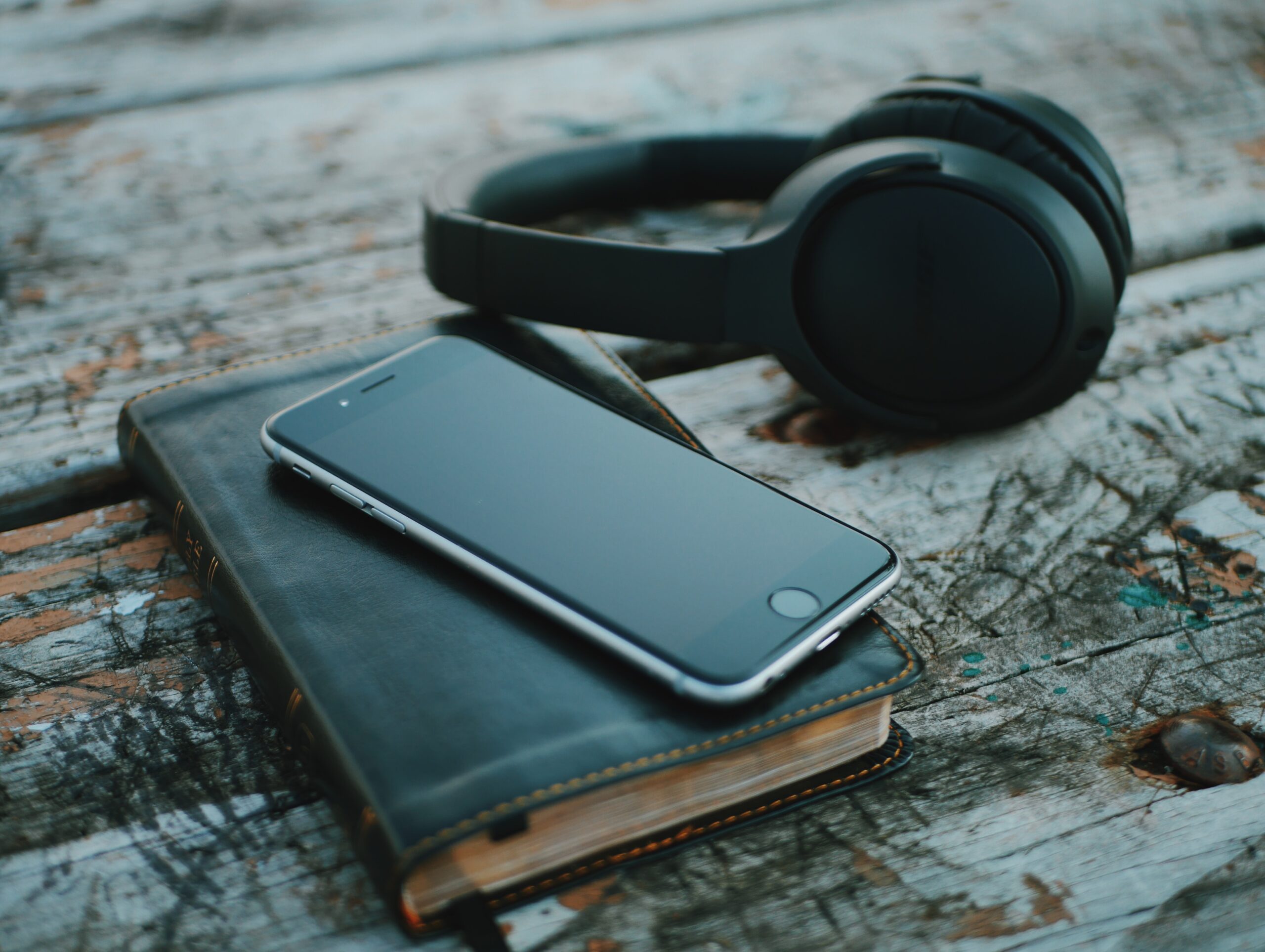 Black smartphone on black diary, over-ear headphones off to the side, on scratched wood slat table. Using as audiobook image